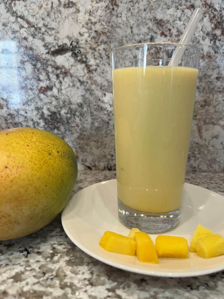 FBB_Beverage_Mango.Bliss.Yogurt.Delight_Hero v2_1200x1600. A Mango Lassi (Will's Version) in a tall glass with a straw on a plate with cubes of mango.