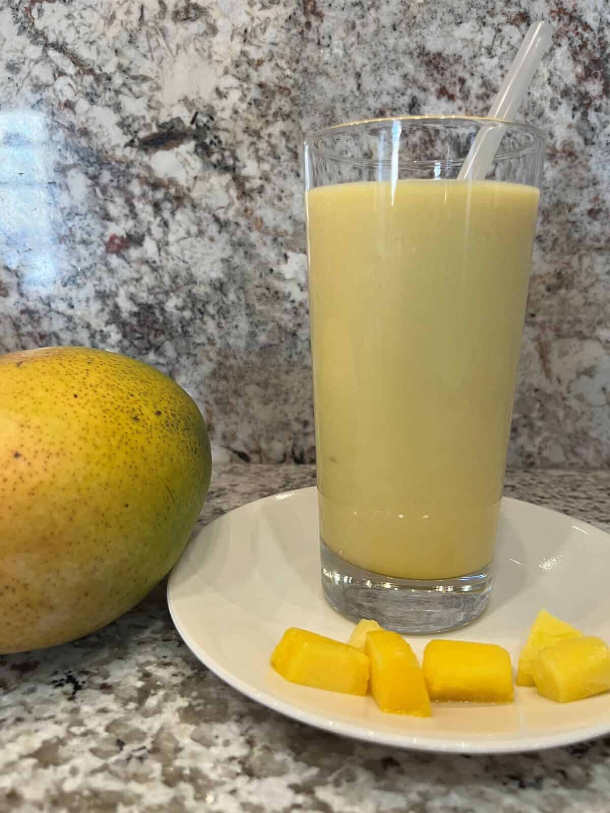 FBB_Beverage_Mango.Bliss.Yogurt.Delight_Hero v2_1200x1600. A Mango Lassi (Will's Version) in a tall glass with a straw on a plate with cubes of mango.