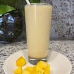 FBB_Beverage_Mango.Bliss.Yogurt.Delight_Hero v2_1200x1600. Hero v2_1200x1600. A Mango Lassi (Will's Version) in a tall glass with a straw on a plate with cubes of mango.