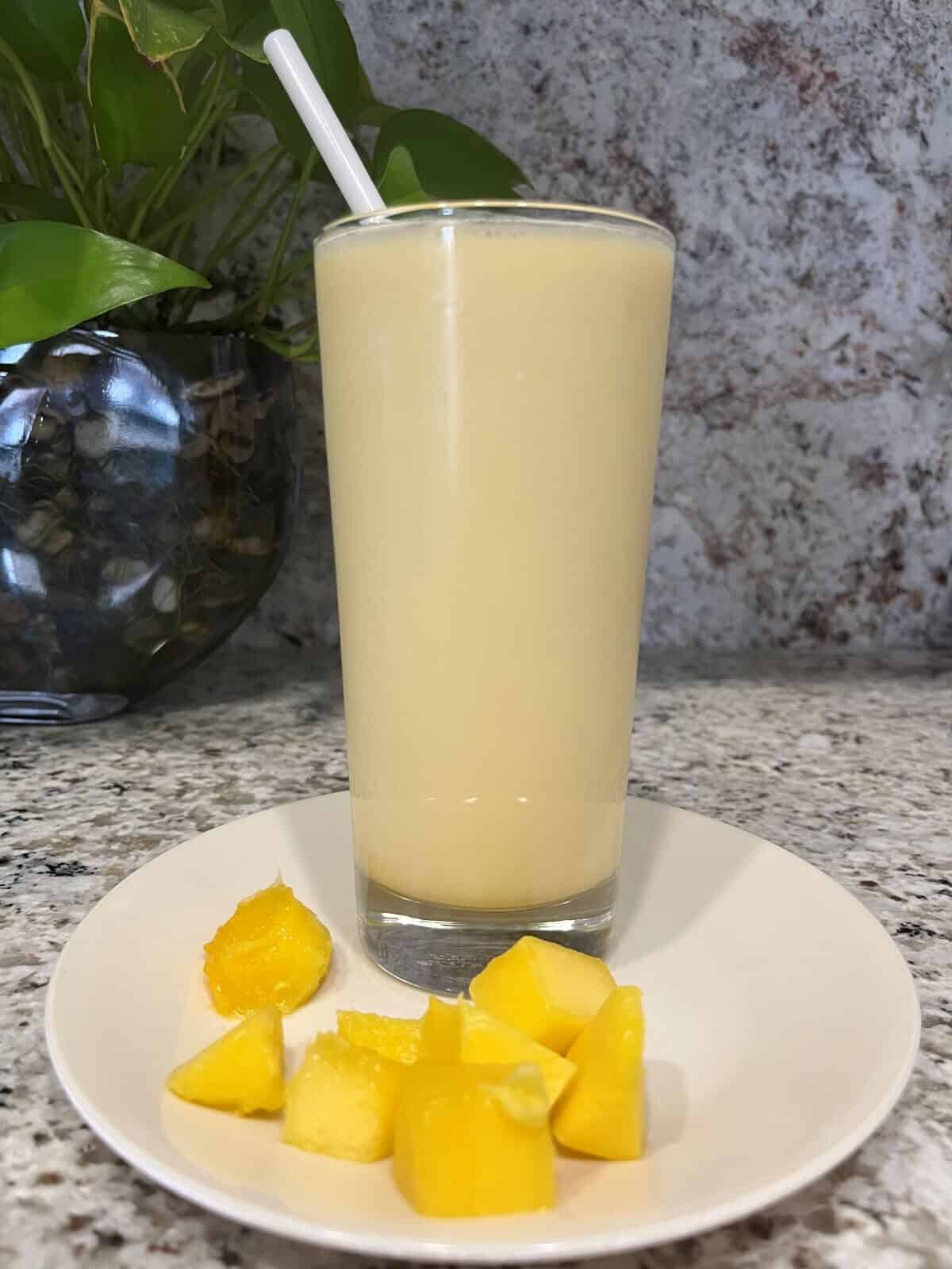 FBB_Beverage_Mango.Bliss.Yogurt.Delight_Hero v2_1200x1600. Hero v2_1200x1600. A Mango Lassi (Will's Version) in a tall glass with a straw on a plate with cubes of mango.