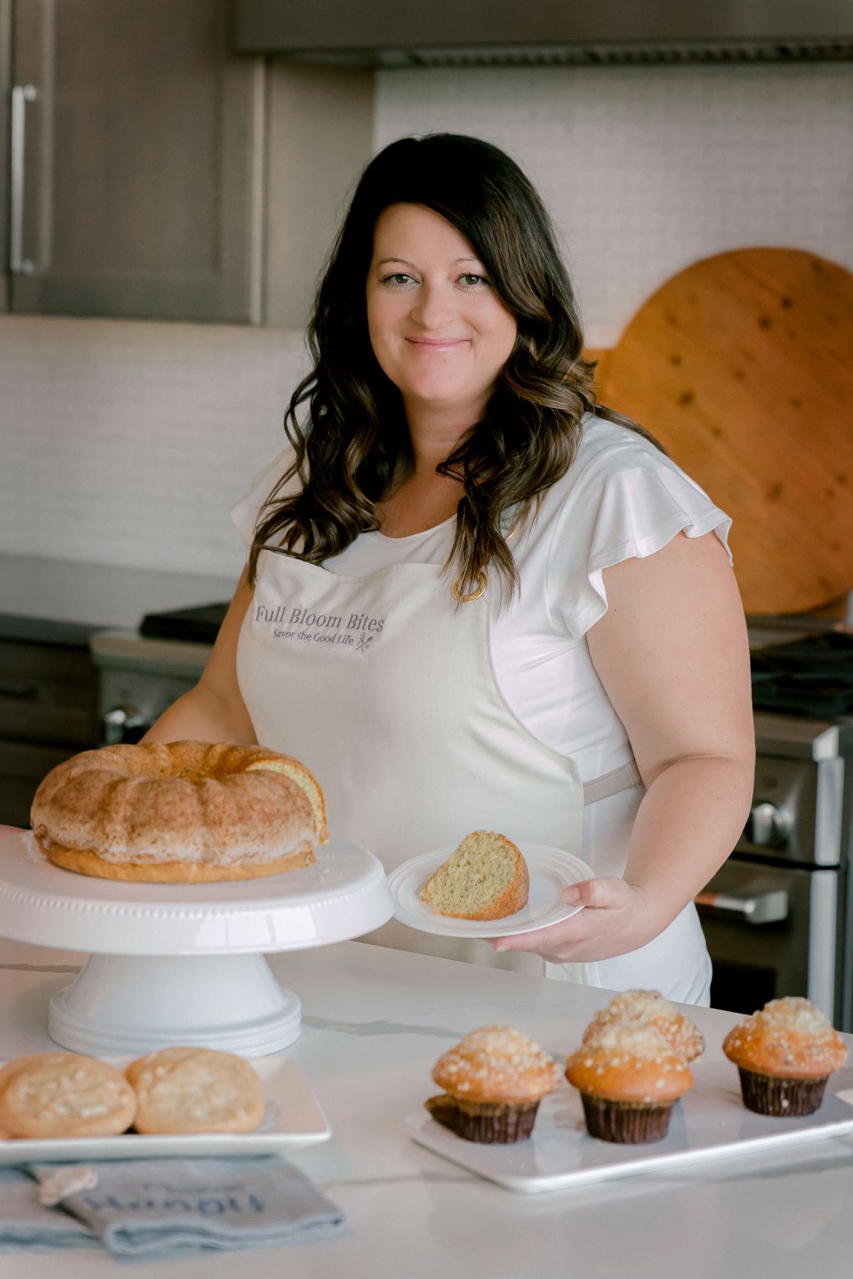 FBB_Jen_Slice.of.Cake_2024. Jen in the kitchen holding a delicious slice of Almond Poppyseed Bunt Cake with a tray of cookies and muffins on display.