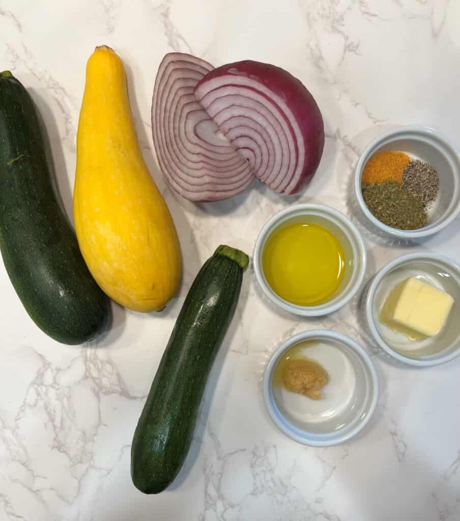 FBB_Side.Zucchini.Squash.Ingredients.1200x1359. This is an ingredient photo of fresh zucchini, yellow squash, red onion, olive oil, butter, garlic and trio of seasonings.