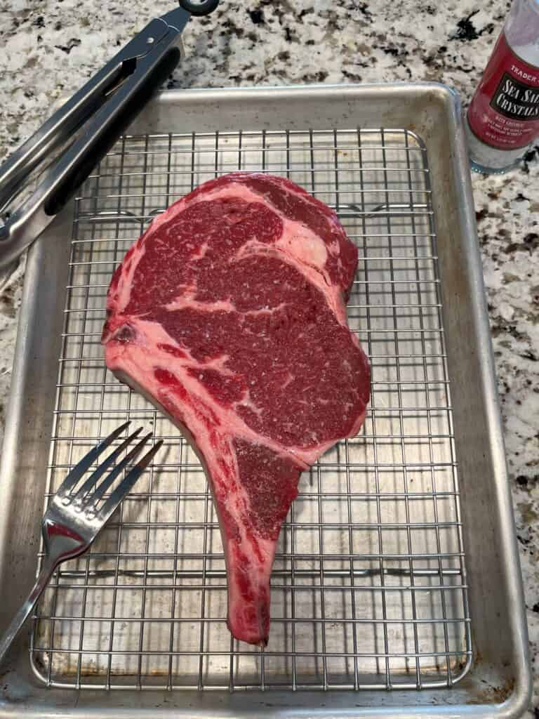 A bone-in ribeye steak being dry brined with a generous coating of sea salt on a half-sheet pan with a grate