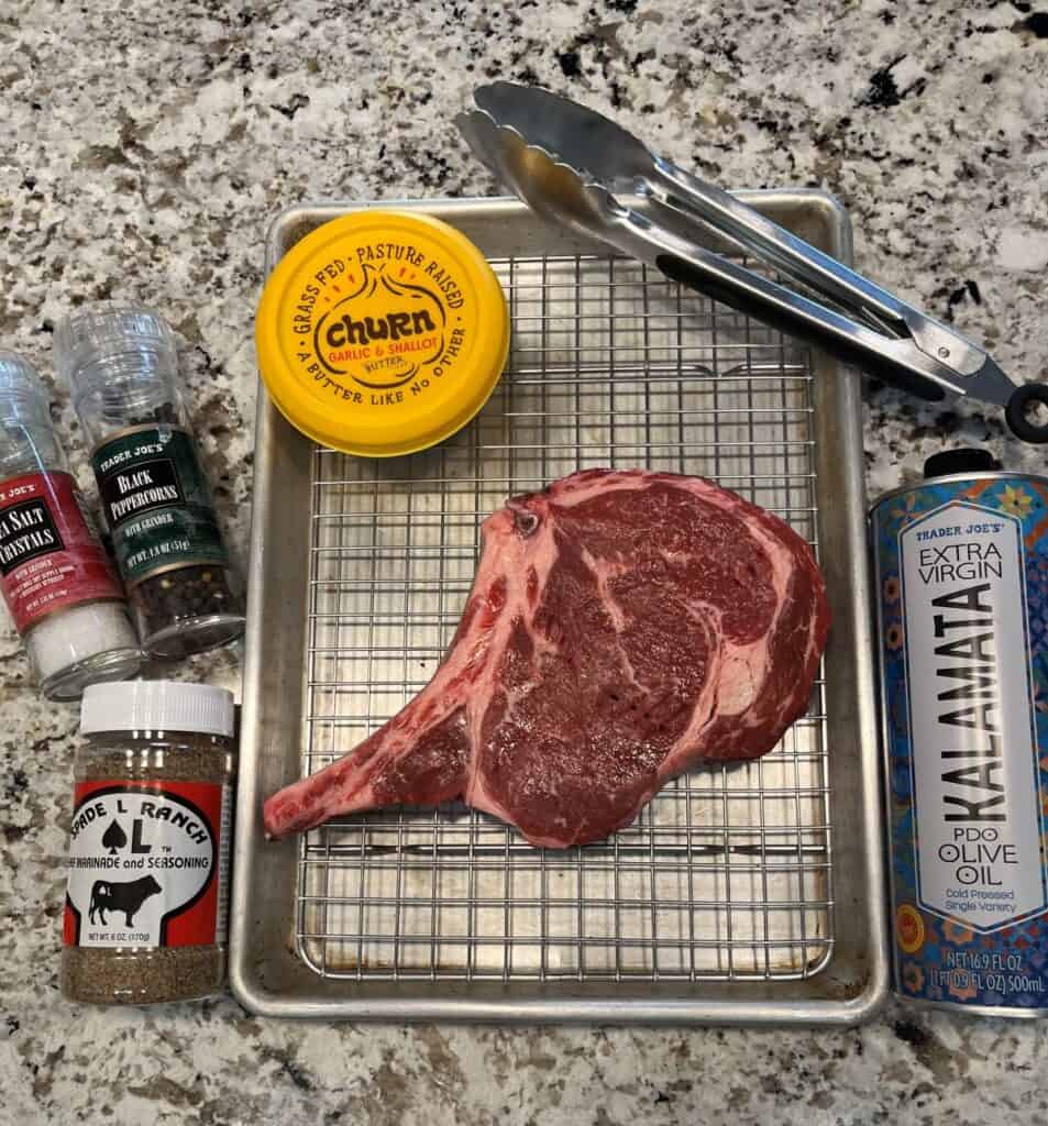 Ingredients for a perfect steak. A Bone-in ribeye steak on a sheet pan with a grate, cold-pressed olive oil, salt, pepper, Spade L Ranch seasoning, and garlic and shallot butter.