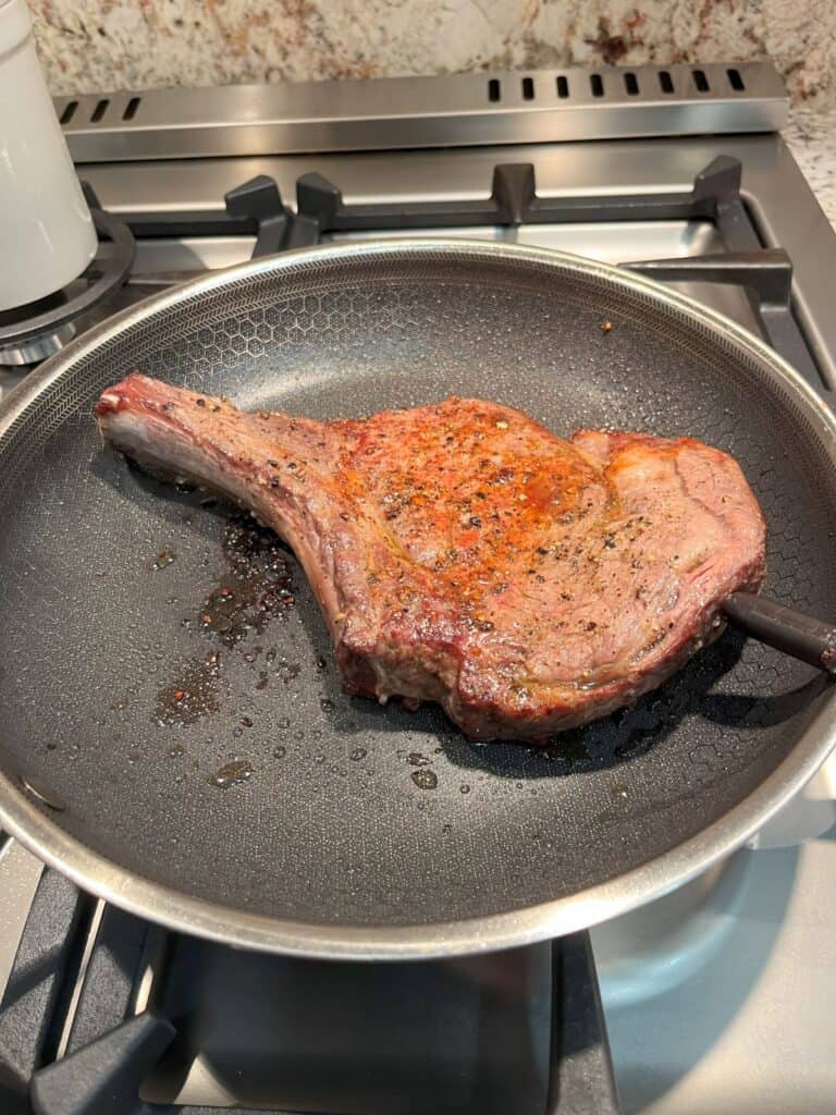 A bone-in ribeye steak sizzling in a HexClad frying pan on the stove, with a Yummly Smart Thermometer inserted to ensure perfect cooking temp.