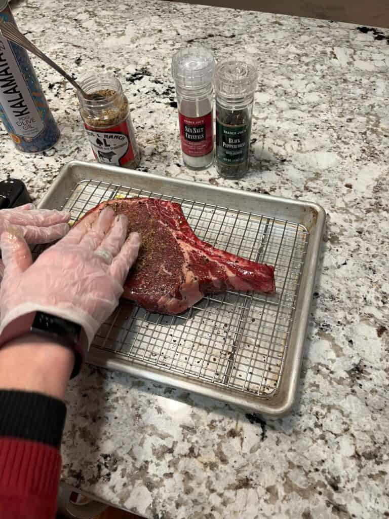 A bone-in ribeye steak on a sheet pan with a grate, seasoned with cold-pressed olive oil, pepper, Spade L Ranch seasoning.