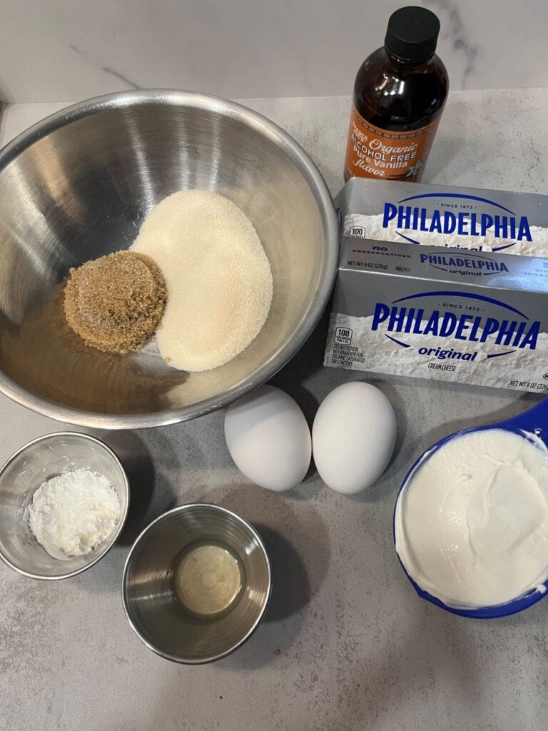 Cheesecake ingredients arranged on a countertop: cream cheese, sugars, sour cream, eggs, vanilla, and cornstarch.