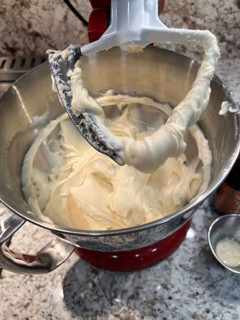 Creamy whipped mixture clinging to a stand mixer paddle above a metal mixing bowl, with vanilla extract nearby.