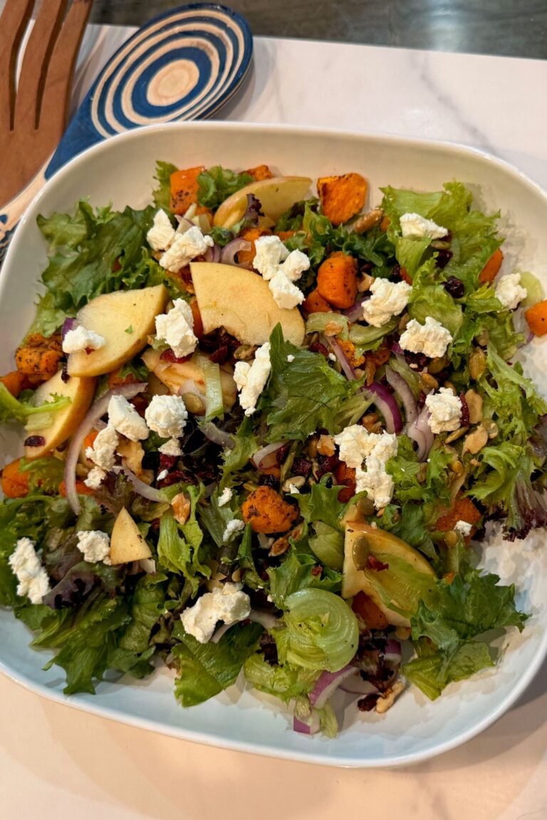 Fall harvest salad on a rimmed plate with roasted squash, apples, greens, goat cheese, nuts, and cranberries.
