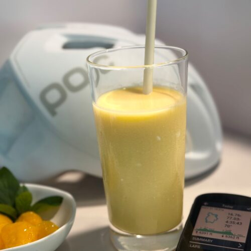 Glass of thick, creamy mango lassi with a straw, sitting on a kitchen counter in front of a blurred cycling helmet