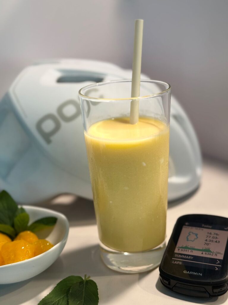 Glass of thick, creamy mango lassi with a straw, sitting on a kitchen counter in front of a blurred cycling helmet