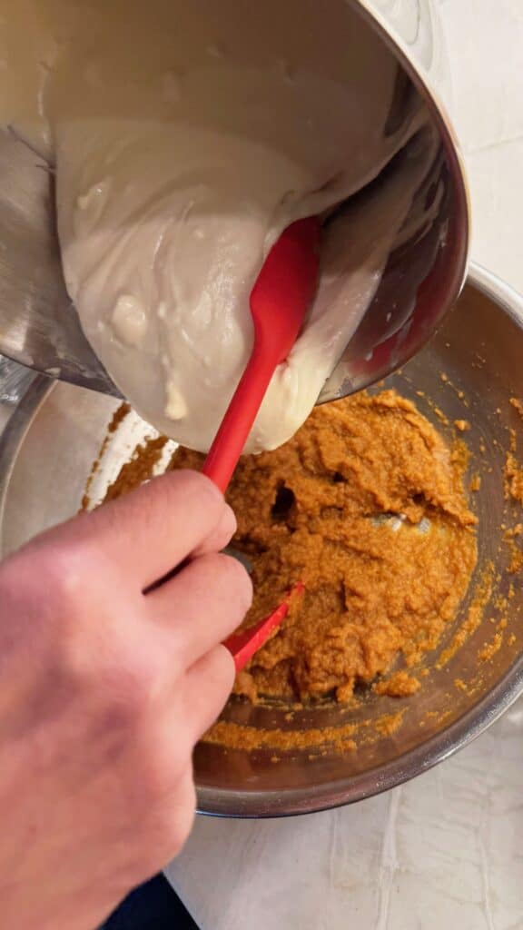 Creamy NY-style cheesecake mixture being poured into a bowl of pumpkin filling with a red spatula.