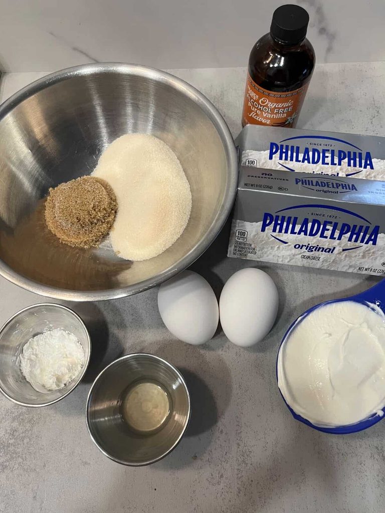 Cheesecake ingredients arranged on a countertop: cream cheese, sugars, sour cream, eggs, vanilla, and cornstarch.