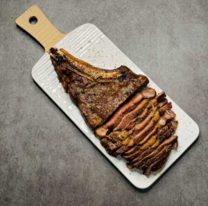 A single cooked bone-in ribeye steak on a white decorative cutting board cut into thin slices.