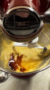Red KitchenAid stand mixer blending batter with visible vanilla extract and maple extract in a stainless steel bowl.