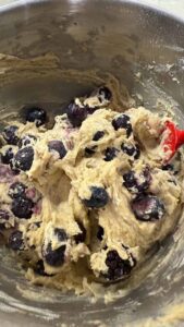 Thick batter with fresh or frozen blueberries being folded with a red spatula in a mixing bowl.