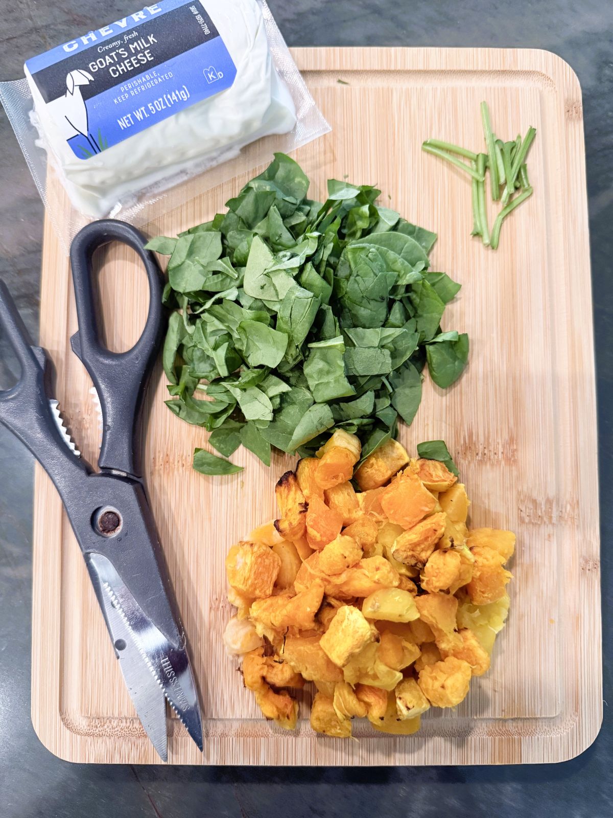 Freshly chopped quiche ingredients on a cutting board, including a package of goats milk cheese, diced squash and spinach with a knife nearby.