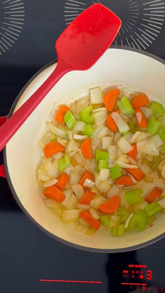 Chopped carrots, celery, and onions sautéing in a white pot with a red spatula on a stovetop.