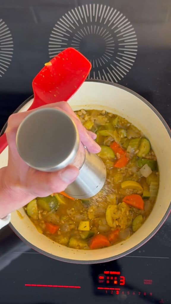 Hand seasoning a pot of vegetable stew with a pepper grinder on a modern stovetop.