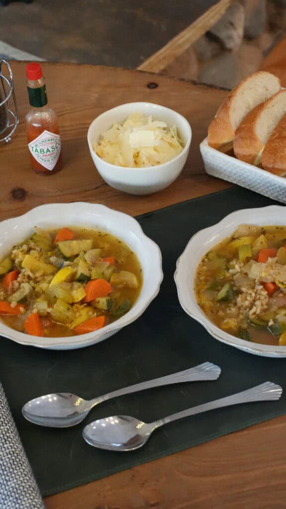 Two bowls of vegetable barley soup on a wooden table, served with cheese, hot sauce, and sliced bread