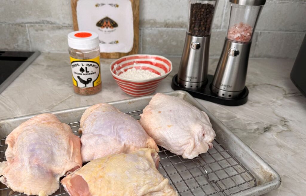 Raw chicken thighs on a wire rack with flour, ranch seasoning, salt, and pepper arranged on a kitchen counter for air‑fryer fried chicken prep.