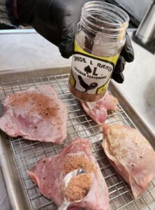 A gloved hand sprinkling chicken seasoning over raw chicken thighs arranged on a wire rack for cooking.