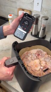 A hand holding a smartphone with air‑fryer settings displayed while placing seasoned chicken pieces into an air‑fryer basket lined with parchment.