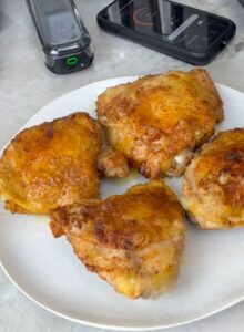 A plate of golden, crispy cooked chicken thighs with a smartphone and meat thermometer nearby for temperature monitoring.