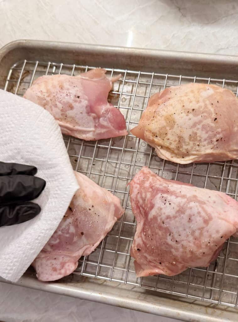 A gloved hand patting raw chicken thighs dry on a wire rack to prepare them for seasoning and cooking.