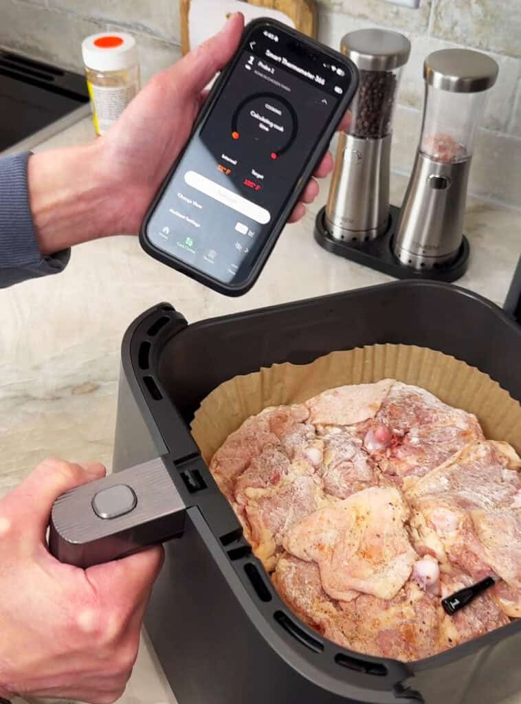 A hand holding a smartphone with air‑fryer settings displayed while placing seasoned chicken pieces into an air‑fryer basket lined with parchment.