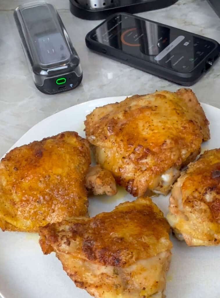 A plate of golden, crispy cooked chicken thighs with a smartphone and meat thermometer nearby for temperature monitoring.