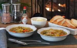 Two bowls of vegetable soup on a wooden table in front of a lit fireplace, served with bread, cheese, and Tabasco.