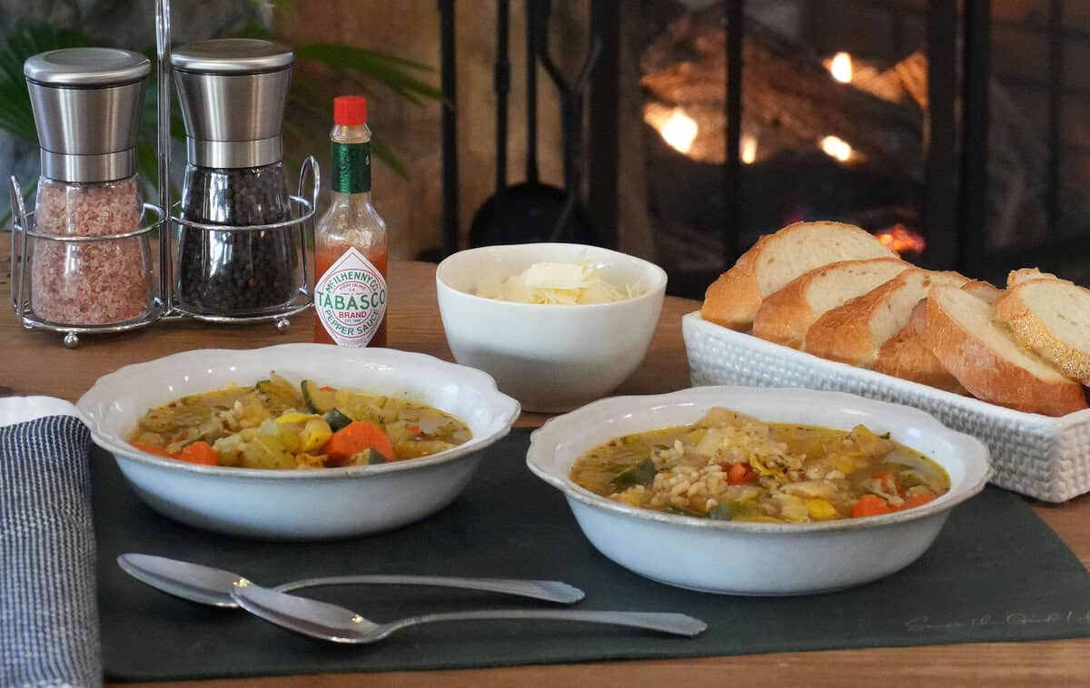 Two bowls of vegetable soup on a wooden table in front of a lit fireplace, served with bread, cheese, and Tabasco.