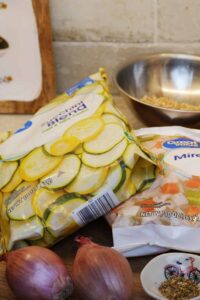 Frozen zucchini and mirepoix bags with shallots, herbs, and grains arranged on a kitchen counter.