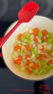 Chopped carrots, celery, and onions sautéing in a white pot with a red spatula on a stovetop.
