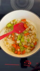 Seasoning with dried herbs into a pot of sautéing carrots, celery, and onions on a hot stovetop.