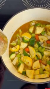 Hand stirring vibrant vegetable soup in a pot on a modern stovetop, filled with zucchini, squash, carrots, celery, and onions.