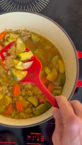 Hand lifting a ladle of vegetable soup from a red-handled pot on a hot stovetop