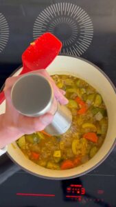 Hand seasoning a pot of vegetable stew with a pepper grinder on a modern stovetop.