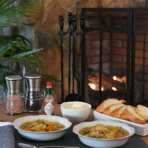 Two bowls of vegetable soup on a wooden table in front of a lit fireplace, served with bread, cheese, and Tabasco.