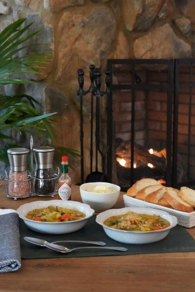 Two bowls of vegetable soup on a wooden table in front of a lit fireplace, served with bread, cheese, and Tabasco.