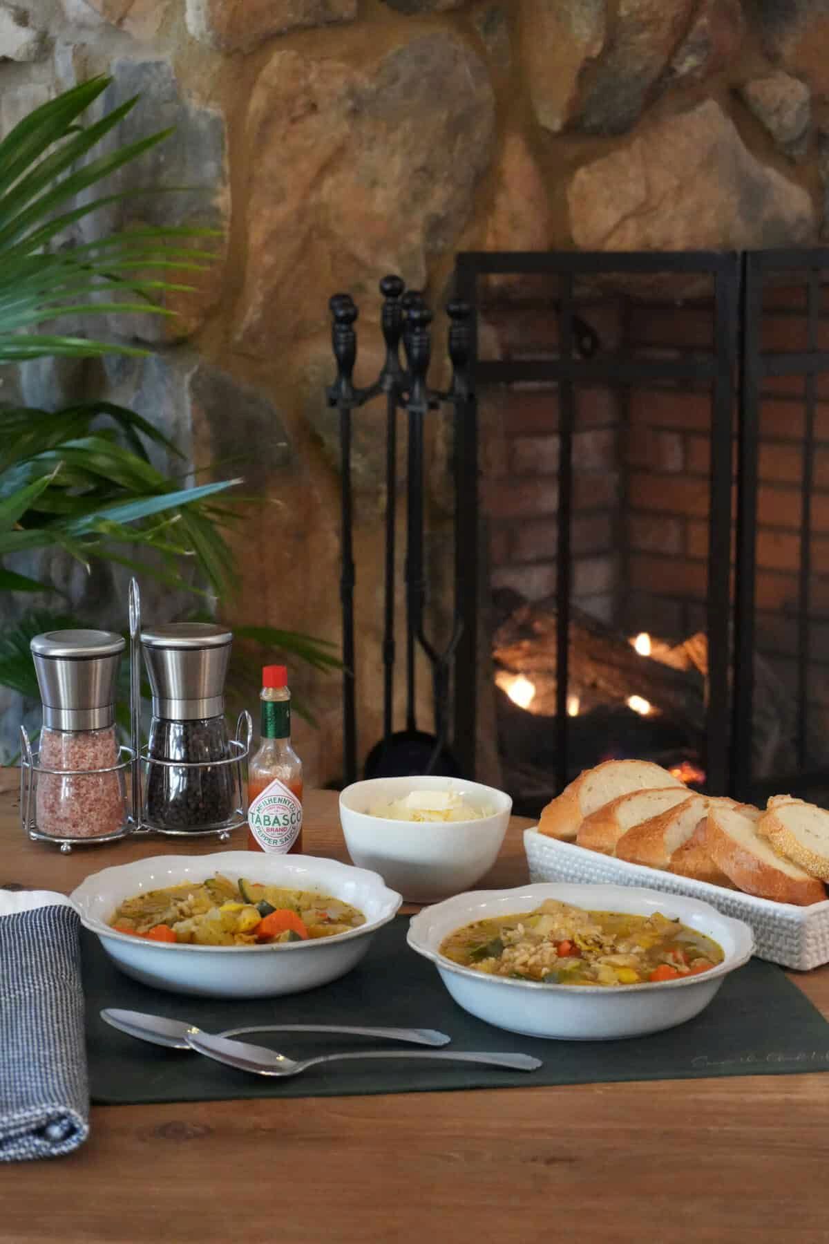 Two bowls of vegetable soup on a wooden table in front of a lit fireplace, served with bread, cheese, and Tabasco.
