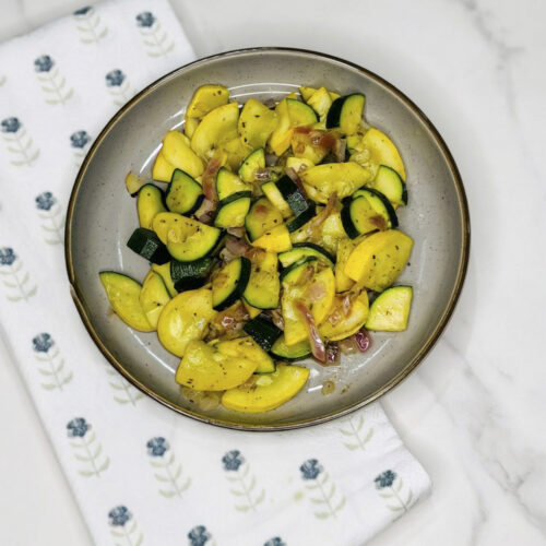 Plate of sautéed zucchini, yellow squash, and red onion served with a fork on a white cloth.