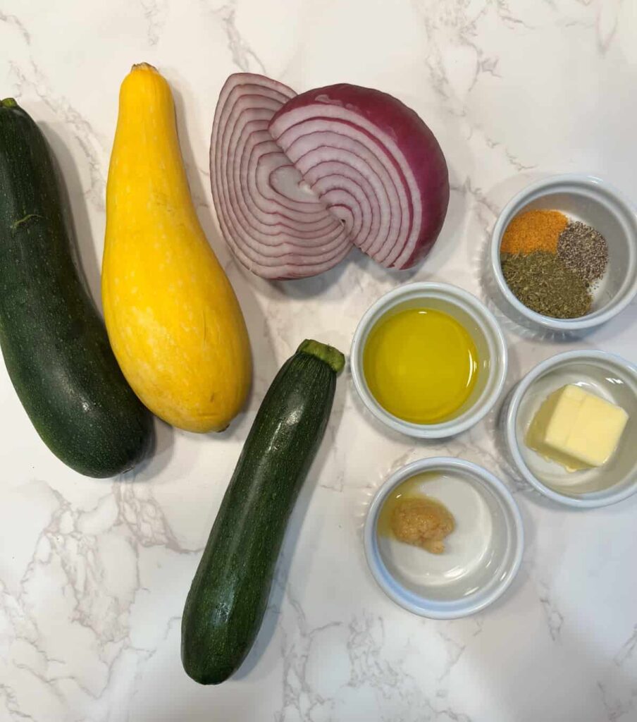Zucchini, yellow squash, olive oil, seasonings, and aromatics arranged on a light surface for a sautéed vegetable recipe.