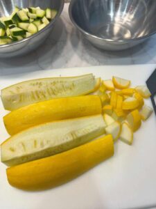 Dicing yellow squash into quarters with already‑cut zucchini in a stainless steel bowl; the empty bowl is prepared for sliced red onion.