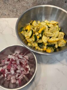 Chopped zucchini and yellow squash in a stainless steel bowl next to a second bowl filled with diced red onion, ready for the next sautéing step.