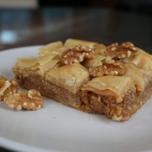 Pieces of baklava on a white plate, topped with walnuts and layered with golden, flaky filo pastry.