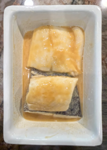 Black cod fillet marinating in a light brown honey miso marinade inside a white rectangular dish.