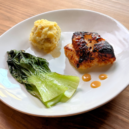 Pan‑seared black cod with a caramelized crust served alongside sautéed greens and a creamy mashed potato scoop on a white plate.