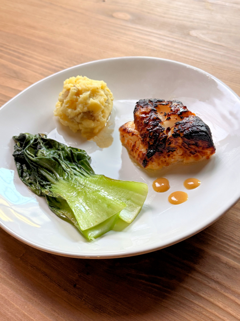 Pan‑seared black cod with a caramelized crust served alongside sautéed greens and a creamy mashed potato scoop on a white plate.